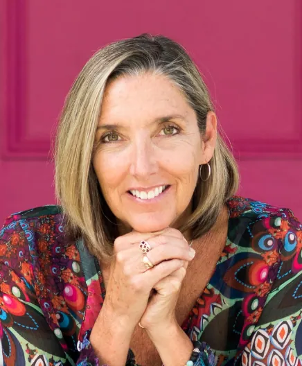Portrait of a woman with shoulder-length blonde hair, smiling with hands clasped, wearing a colorful patterned top