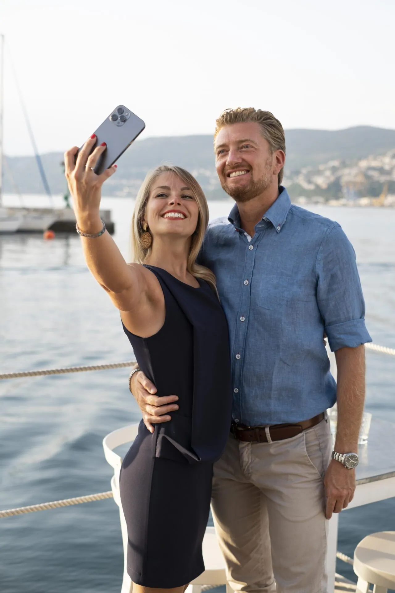 Couple taking a smiling selfie together on a boat deck at a Mediterranean marina at golden hour