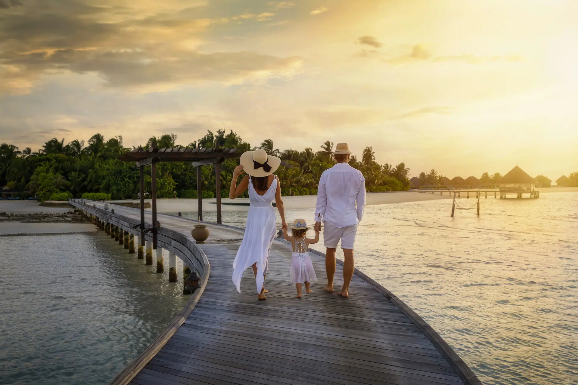 Family of three walking hand in hand along an overwater bungalow boardwalk at sunset