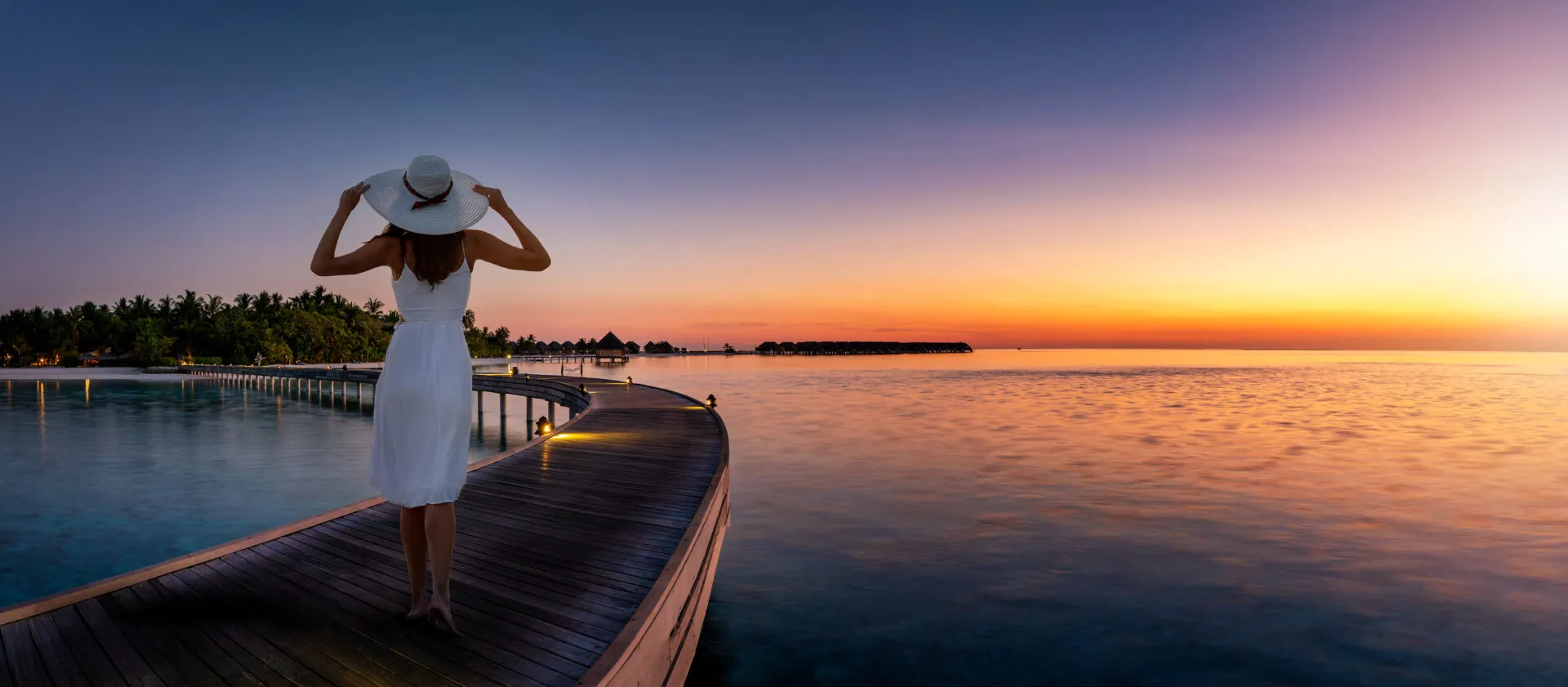 Woman in a white dress watching a vivid sunset from an overwater boardwalk