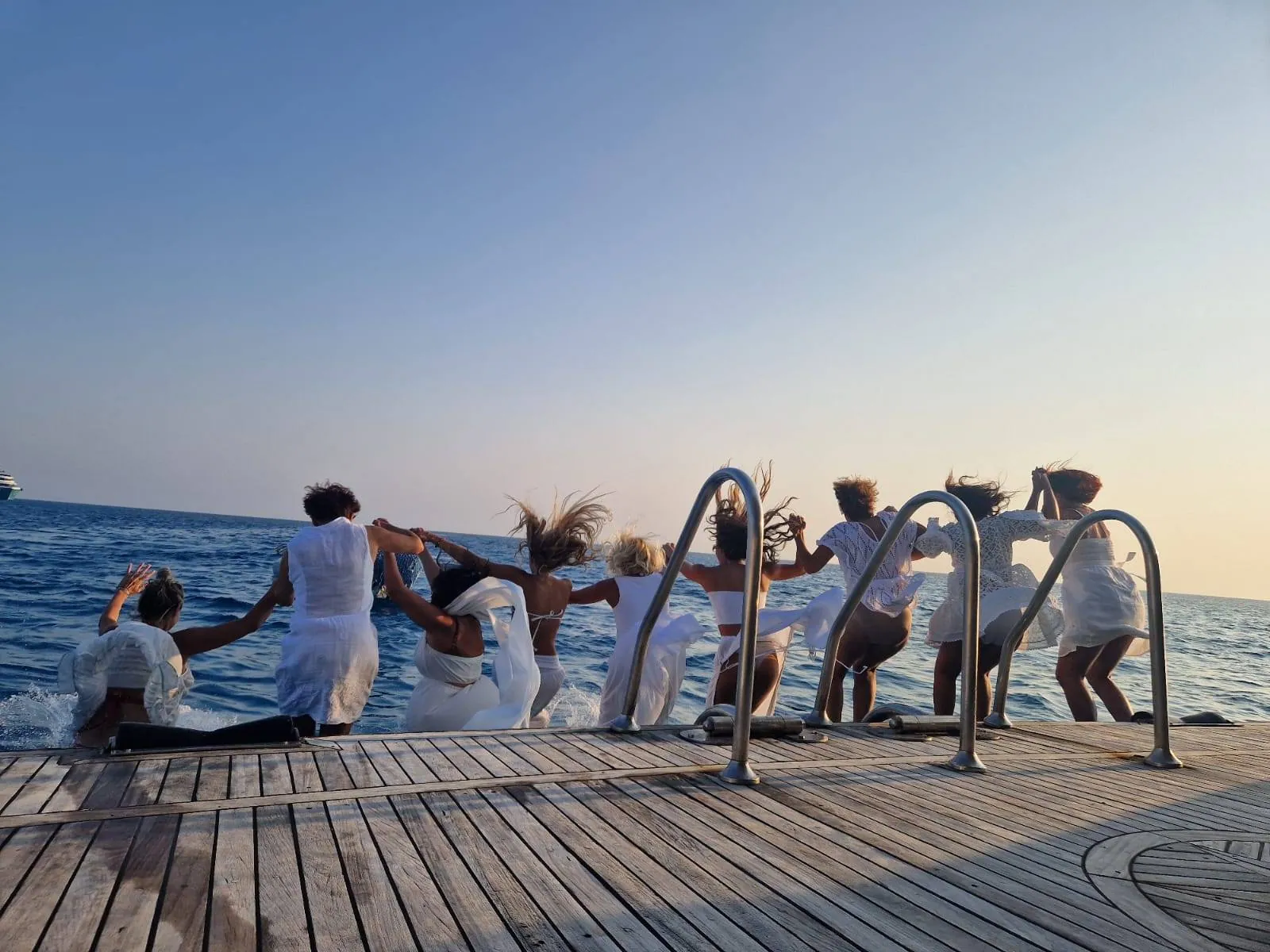 Group of women in white joyfully holding hands on a yacht's swim platform at sunset