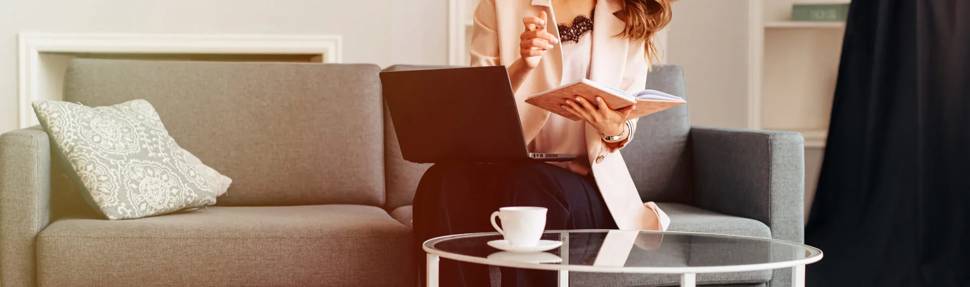 Woman reading on a sofa beside a laptop and coffee cup in a bright room