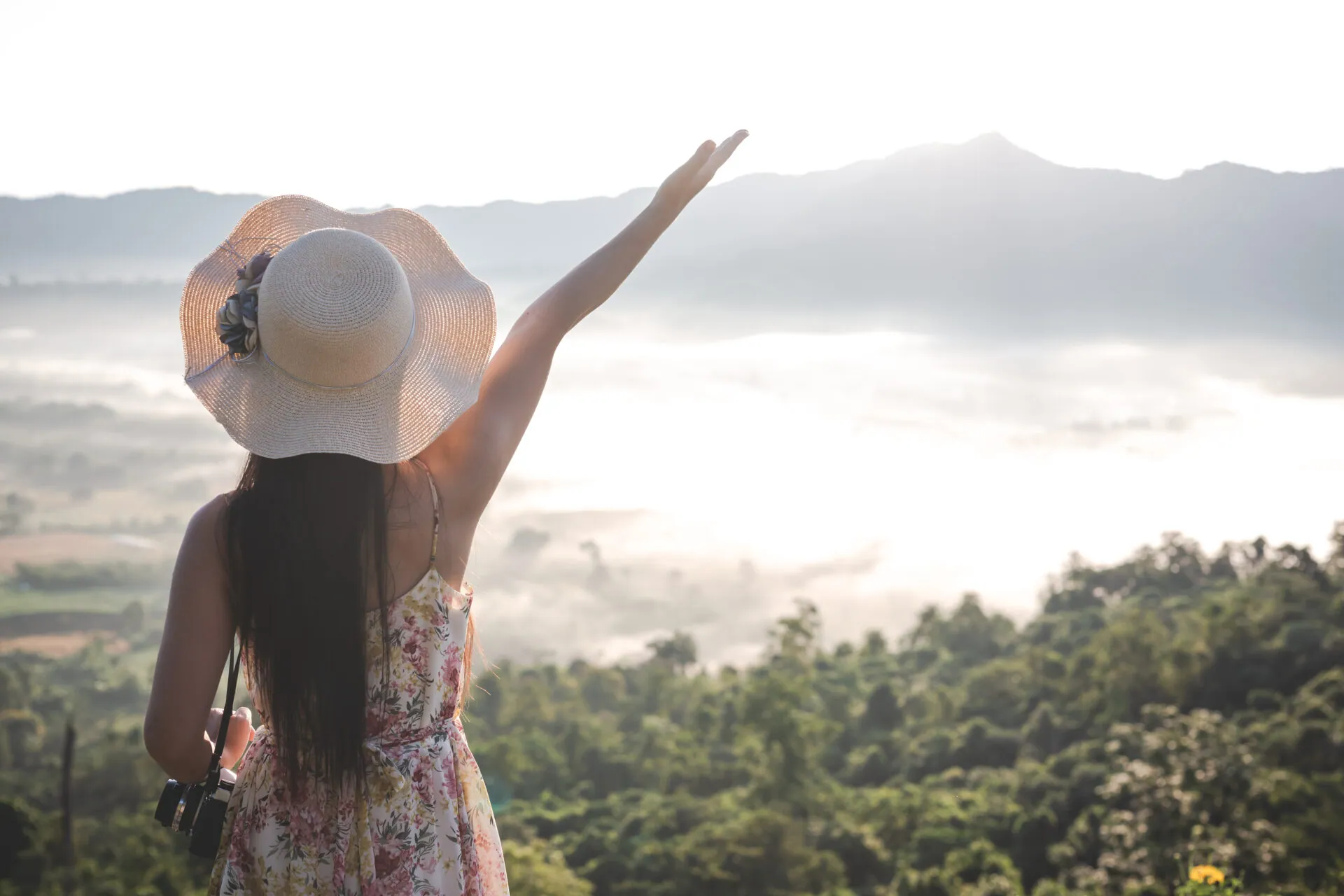 Woman in a sun hat raising her arm above misty forested mountains